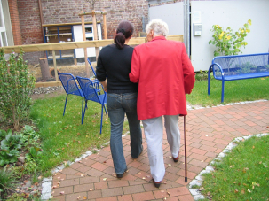 Photo d'un personne âgée de dos accompagnée par une aidante dans un jardinet