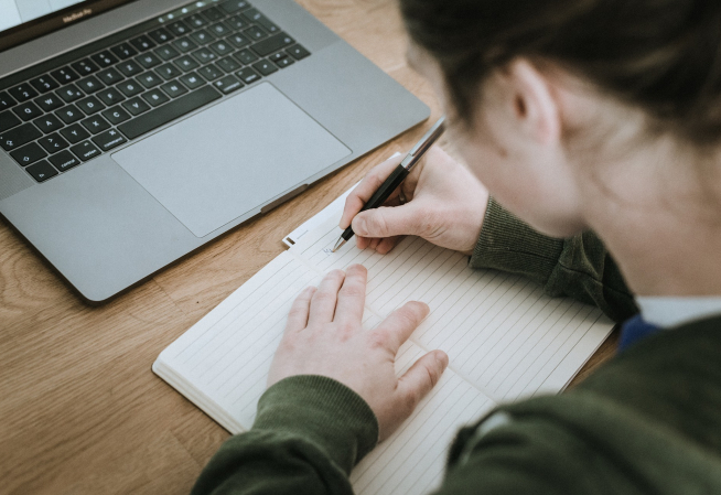 Photo d'une personne jeune, de dos, devant un écran d'ordinateur, en train de prendre des notes sur une feuille de papier