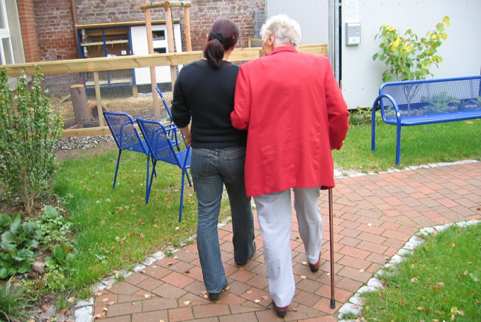 Photo d'un personne âgée de dos accompagnée par une aidante dans un jardinet